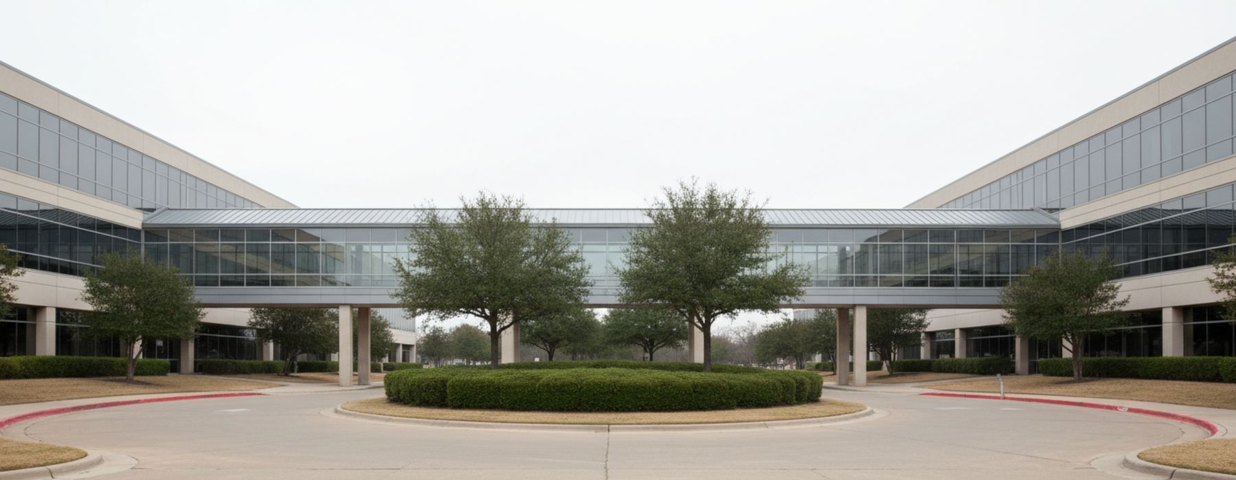 Office campus with two glass buildings joined by a covered walkway on slim columns, a circular drive with a clipped shrub island and oaks under a white sky.